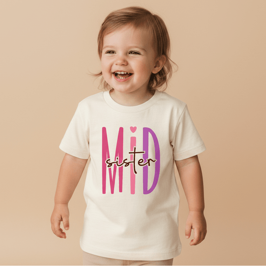 A smiling toddler wears the Toodleroos Middle Sister Toddler Shirt – Bold Sibling Tee, featuring “Mid Sister” in playful pink and purple lettering on a classic white shirt, standing against a beige background—an ideal pick for matching sister shirts or sibling collections.