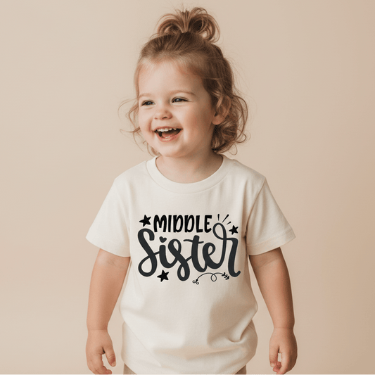 A smiling young child with light brown hair in a ponytail wears the Toodleroos Middle Sister Toddler Shirt, featuring a playful hand-lettered sibling design in black on white—an ideal gift for siblings. She stands against a beige background.