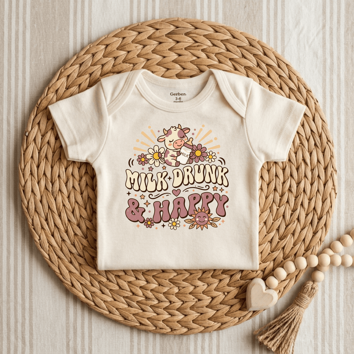 A cream Milk Drunk & Happy Onesie – Cute Baby Cow Bodysuit by Toodleroos is displayed on a woven mat, adorned with a whimsical cartoon cow surrounded by flowers and the cheerful phrase “Milk Drunk & Happy” in playful lettering. Perfect for fans of animal-themed baby apparel, this adorable piece adds fun to any little one’s wardrobe. Wooden teether beads and a heart shape nearby make it an ideal baby shower gift.
