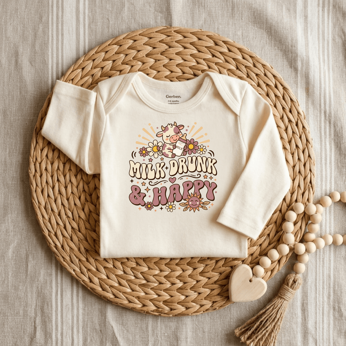 The Milk Drunk & Happy Onesie – Cute Baby Cow Bodysuit by Toodleroos is a cream-colored onesie featuring an adorable cow and floral design with the phrase “Milk-Drunk & Happy.” Displayed on a woven placemat with a wooden bead garland and heart atop a striped fabric background, this animal-themed bodysuit makes the perfect funny baby shower gift for any little one.