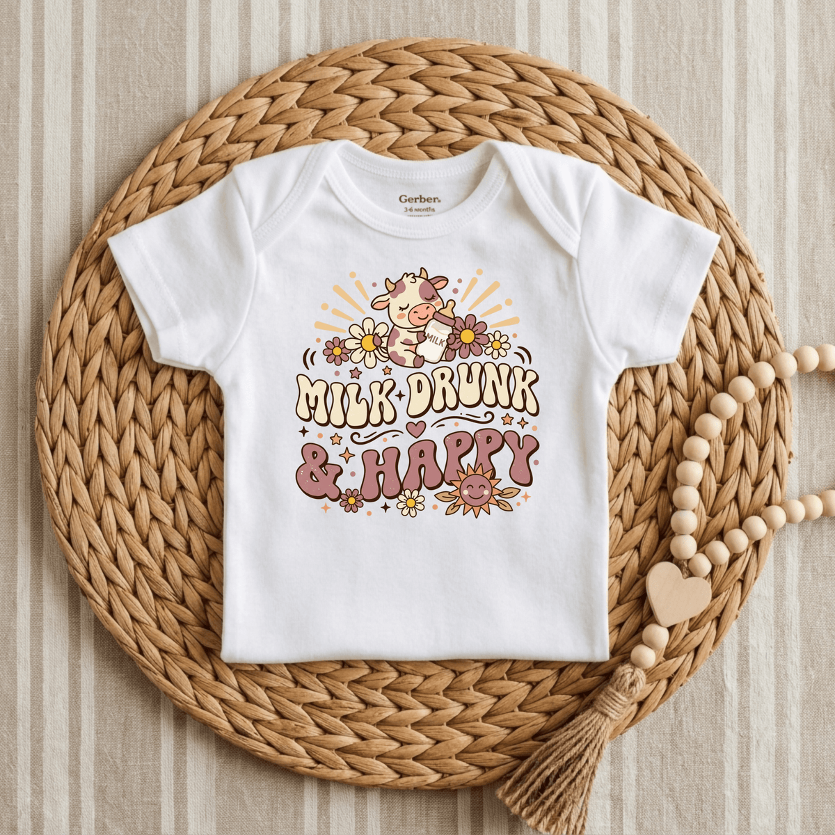 The Milk Drunk & Happy Onesie – Cute Baby Cow Bodysuit by Toodleroos features an adorable illustration of a cow surrounded by flowers and the words “Milk-Drunk & Happy.” Photographed lying on a woven mat beside a beaded wooden garland with a heart-shaped accent, this animal-themed onesie is the perfect funny baby outfit and makes a delightful baby shower gift.
