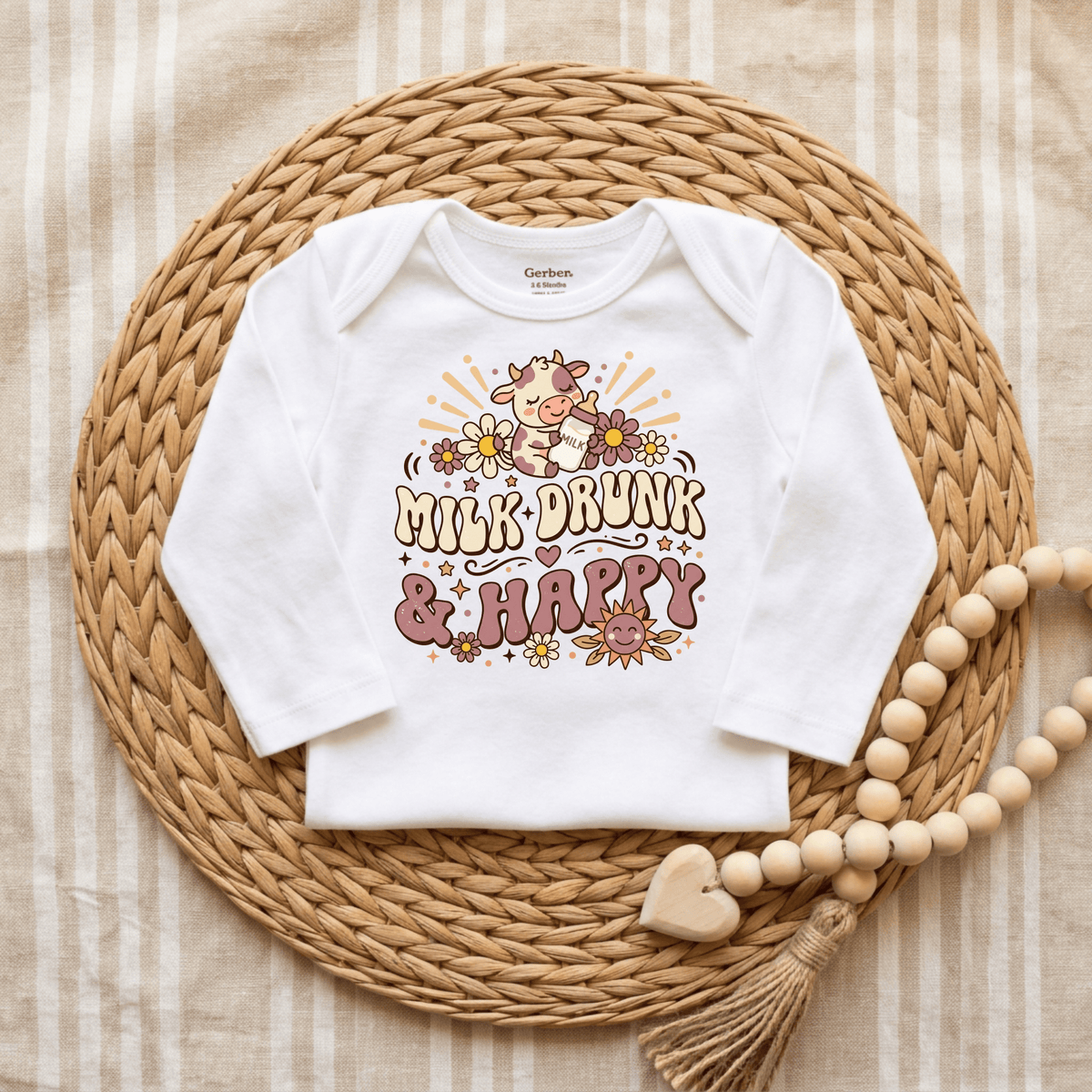 A Toodleroos Milk Drunk & Happy Onesie – Cute Baby Cow Bodysuit is displayed on a woven mat with wooden beads and a heart tassel to the side, atop a striped fabric background—making it one of the cutest animal-themed baby gifts and an ideal choice for baby showers.