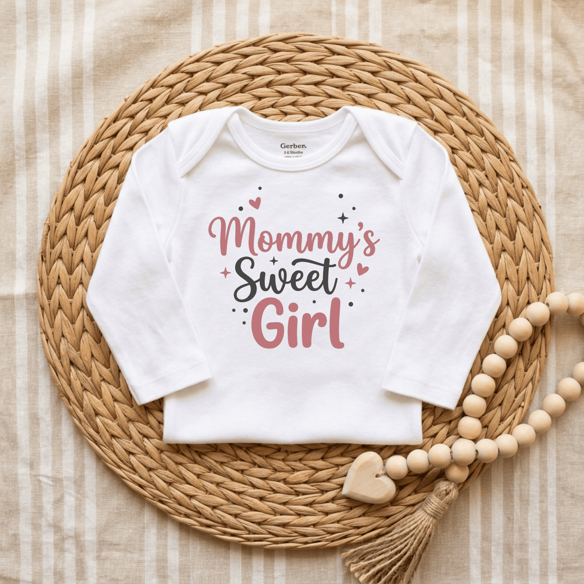 A Toodleroos Mommy’s Sweet Girl Onesie – Baby Girl Long Sleeve Bodysuit in white, featuring “Mommy’s Sweet Girl” in pink and black lettering, is displayed on a woven placemat with a beaded tassel accessory beside it atop a striped fabric background—making it the perfect newborn coming-home outfit.