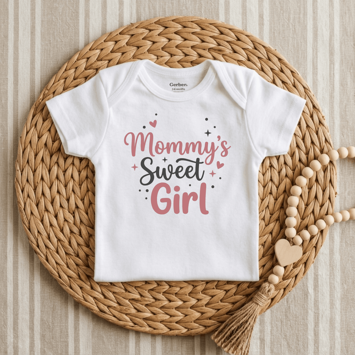 The Toodleroos Mommy’s Sweet Girl Onesie – a baby girl long sleeve bodysuit in white featuring “Mommys Sweet Girl” in pink and black letters, surrounded by hearts and stars – is displayed on a woven mat beside a wooden bead garland with a heart, making it the perfect newborn coming-home outfit.
