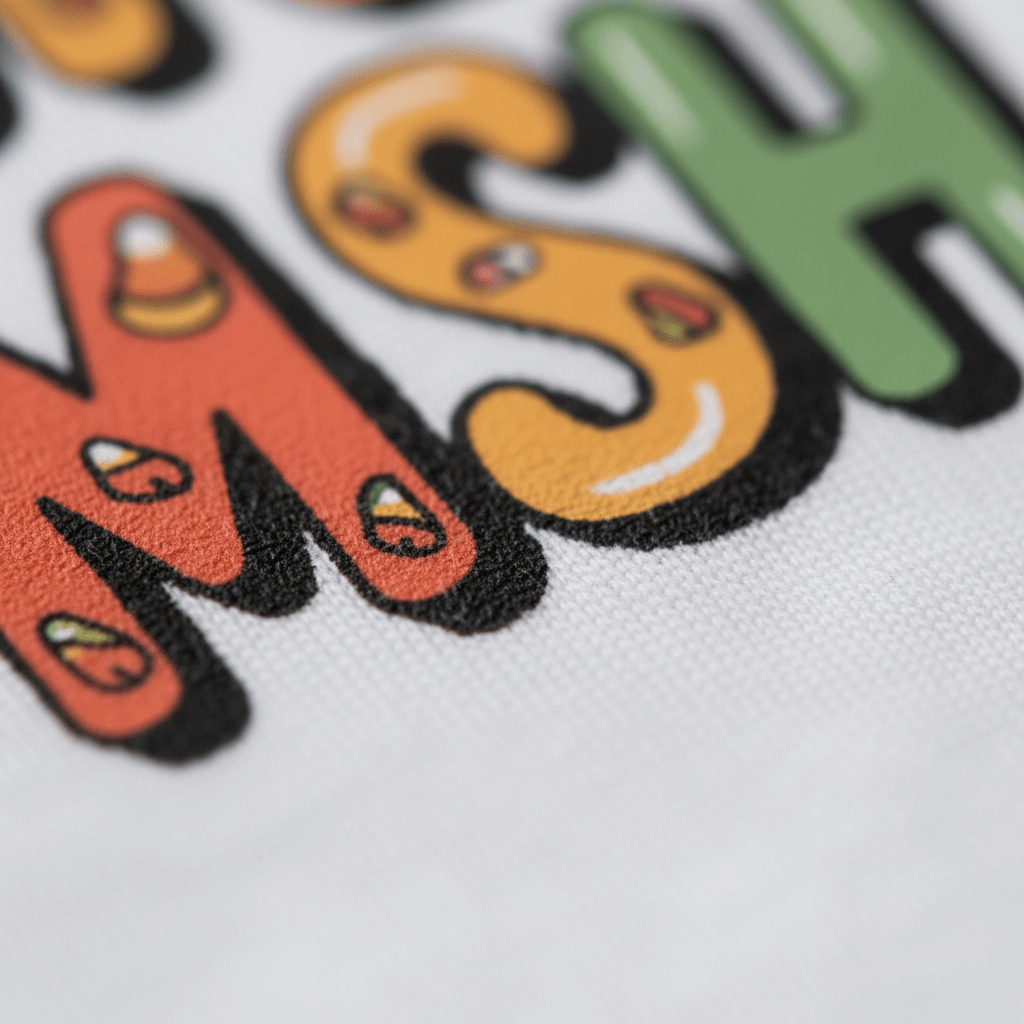 Close-up of colorful letters M and S on fabric, decorated with small candy corn illustrations, from the Toodleroos Monster Mash Bash Halloween Toddler Shirt – a friendly monster tee that’s perfect for a toddler Halloween shirt or kids’ Halloween party shirt.