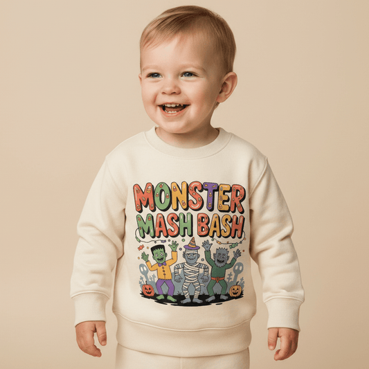 Smiling toddler wearing a cream sweatshirt with colorful cartoon monsters and the text Monster Mash Bash printed on the front, standing against a plain beige background.