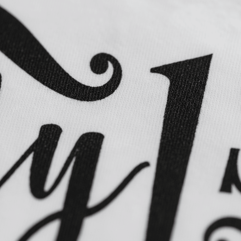 Close-up of black script and bold printed letters on white fabric, likely part of a larger design featured on the Toodleroos “My 1st Christmas Baby Onesie – Holiday Keepsake Bodysuit.” The visible curly “y” and tall “J” suggest festive lettering commonly found on Christmas baby bodysuits.