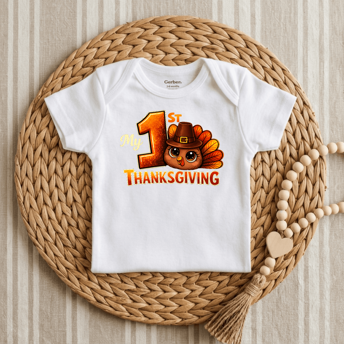 A white Toodleroos My 1st Thanksgiving Onesie – Baby Turkey Bodysuit is displayed on a woven placemat, featuring “My 1st Thanksgiving” and an adorable turkey in a hat printed on the front. Beside the Gerber bodysuit lies a beaded garland with a heart and tassel, making this festive My 1st Thanksgiving onesie the perfect Thanksgiving baby outfit for your little one’s special day.