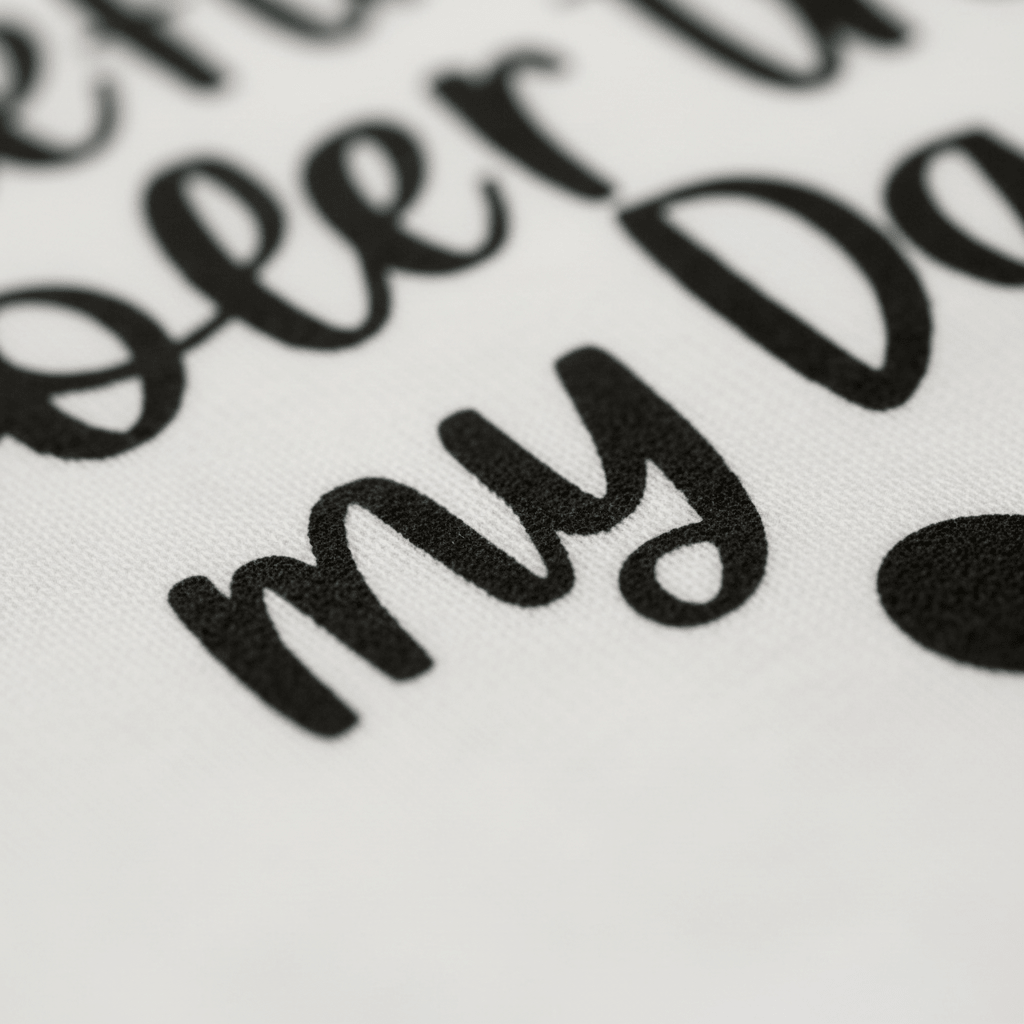 Close-up of black handwritten-style text printed on white fabric, featuring the words “my Da” from the Toodleroos “My Aunt Is Definitely Cooler Than My Dad Toddler Shirt – Funny Family Tee.” The rest of the message is partially visible and blurred, drawing attention to the fabric texture and playful lettering—an ideal cool aunt tee or funny aunt gift, especially as an adorable toddler shirt.