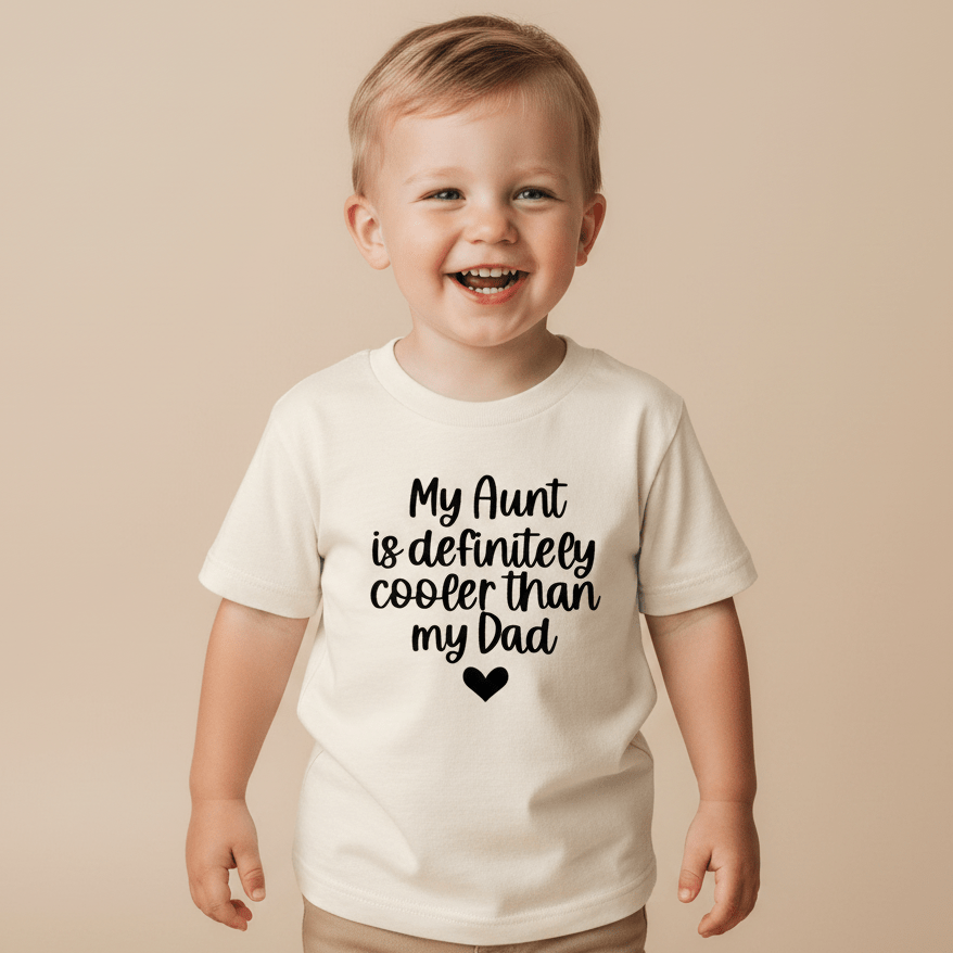 A smiling young child stands wearing the Toodleroos “My Aunt Is Definitely Cooler Than My Dad Toddler Shirt”—a white, funny family tee featuring the playful slogan and a small heart graphic, making it the perfect aunt gift for any fun-loving family.