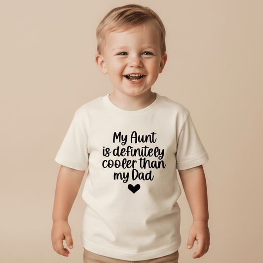 A smiling young child stands wearing the Toodleroos “My Aunt Is Definitely Cooler Than My Dad Toddler Shirt”—a white, funny family tee featuring the playful slogan and a small heart graphic, making it the perfect aunt gift for any fun-loving family.
