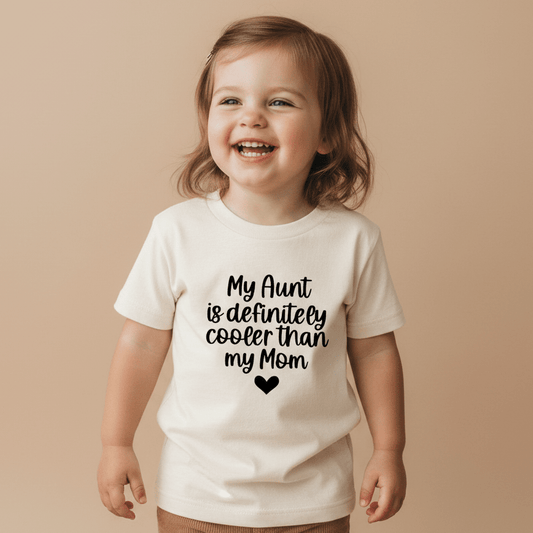 A smiling young child wears the Toodleroos “My Aunt Is Definitely Cooler Than My Mom Toddler Shirt”—a white tee featuring bold black text—while standing against a beige background. This funny family tee is the perfect aunt gift for any occasion.