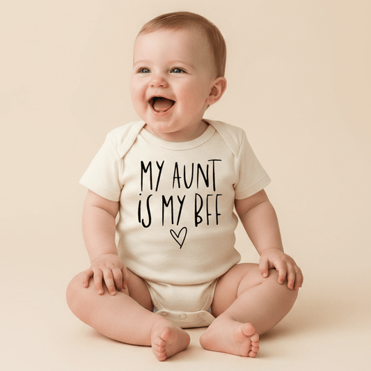 A smiling baby sits in a cream-colored Toodleroos “My Aunt Is My BFF Baby Onesie – Handwritten Aunt Bodysuit,” featuring black text that reads “My Aunt Is My BFF” with a small heart underneath—making it an adorable gift for any aunt.