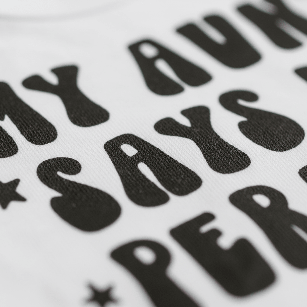A close-up of the Toodleroos My Aunt Says I’m Perfect Baby Onesie – Funny Aunt Bodysuit shows white fabric featuring bold, playful black lettering that reads “MY AUNT SAYS PER,” accented by charming stars. This adorable onesie is a perfect baby shower gift from an aunt, celebrating the unique bond between aunt and baby.