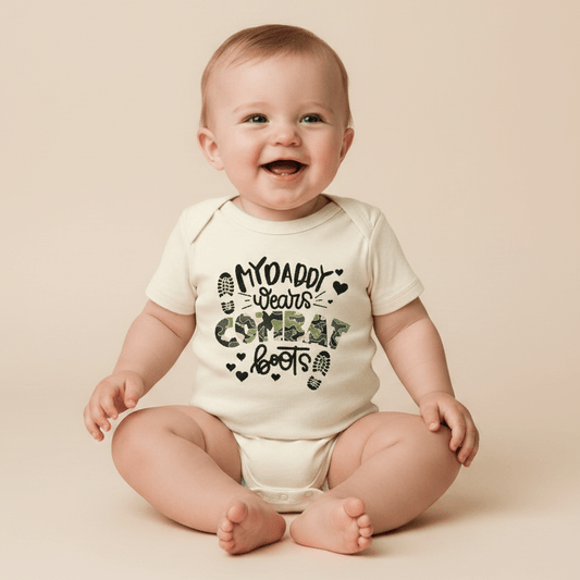 A smiling baby wears the Toodleroos “My Daddy Wears Combat Boots Baby Onesie – Camo Design,” featuring the text “My Daddy Wears Combat Boots” and two boot prints on the front, while sitting against a neutral background—a perfect gift for a military dad.