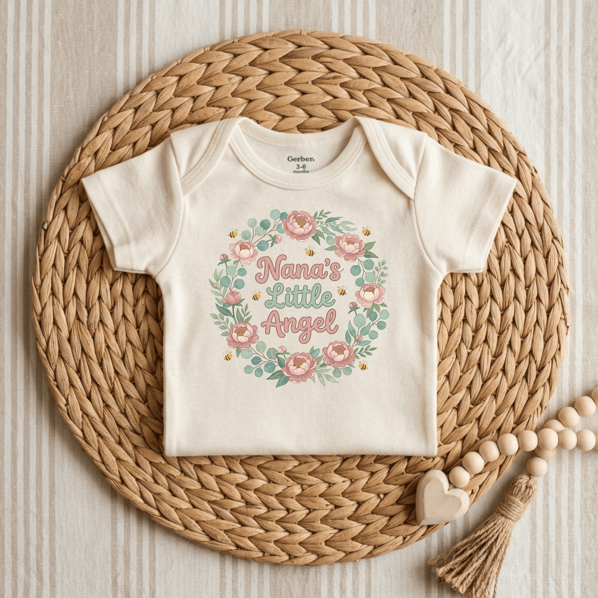 The Toodleroos Nana’s Little Angel Baby Onesie – Floral Grandma Bodysuit is displayed on a woven placemat with a floral wreath and the phrase “Nanas Little Angel.” Wooden beads and heart-shaped decor sit beside it against a striped fabric background. This charming Gerber short sleeve bodysuit is the perfect baby shower gift from Nana.