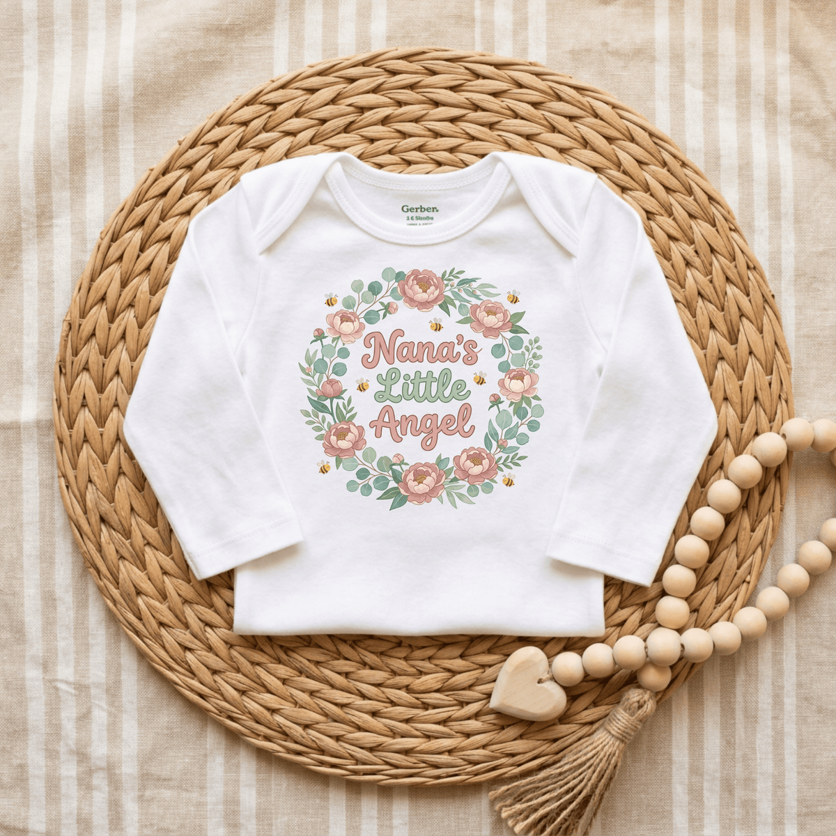 The Toodleroos “Nana’s Little Angel Baby Onesie – Floral Grandma Bodysuit” showcases a charming pink and green floral design on a soft white Gerber short sleeve bodysuit, beautifully styled atop a woven mat with a wooden beaded garland, against a beige striped fabric backdrop—an ideal baby shower gift from Nana.