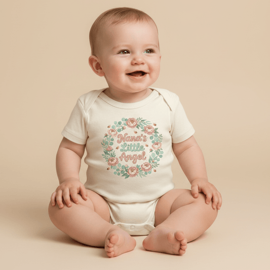 A smiling baby sits on a beige floor, dressed in the Toodleroos Nana’s Little Angel Baby Onesie – Floral Grandma Bodysuit, featuring a pastel floral wreath and sweet lettering—an adorable baby shower gift from Nana.