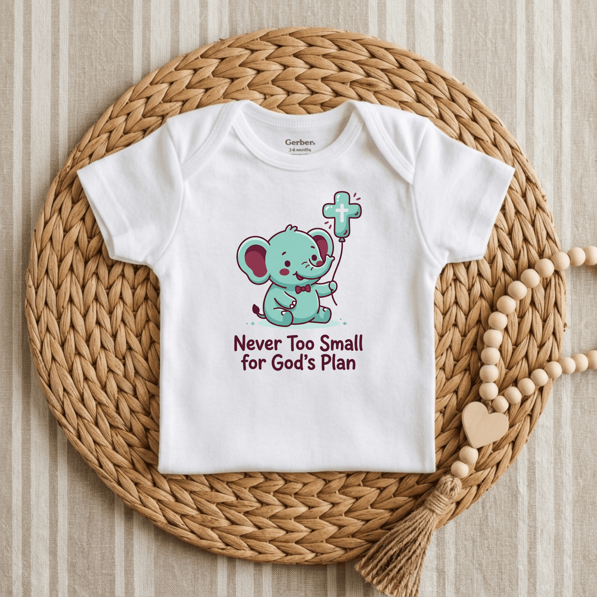 The Toodleroos Never Too Small for God’s Plan Onesie – Faith Bodysuit features a charming blue elephant holding a cross above the inspirational text “Never Too Small for God’s Plan.” This Christian baby onesie, ideal as faith-inspired clothing or a thoughtful baptism gift, is beautifully displayed on a round wicker placemat with a wooden beaded garland beside it.