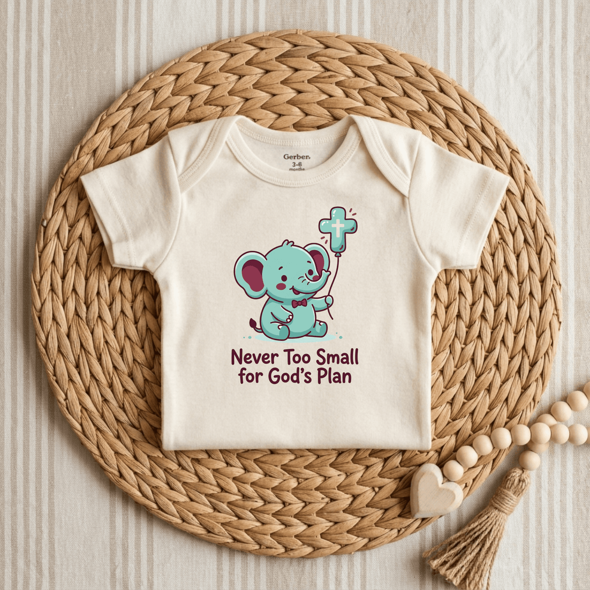 The Toodleroos “Never Too Small for God’s Plan Onesie – Faith Bodysuit” features a cream-colored design with an adorable blue elephant holding a cross above the inspirational text, “Never too small for God’s plan.” Perfect as a Christian baby onesie or baptism gift, this faith-inspired piece is displayed on a round woven mat alongside a wooden bead and heart garland.