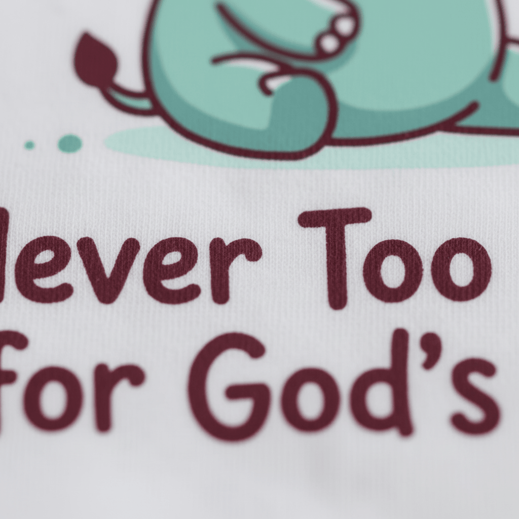 A partial illustration of a green animal, possibly a cartoon dinosaur, appears above the text “Never Too Small for God’s Plan” on white fabric. The Toodleroos Never Too Small for God’s Plan Onesie – Faith Bodysuit is perfect as a baptism gift or Christian baby onesie.