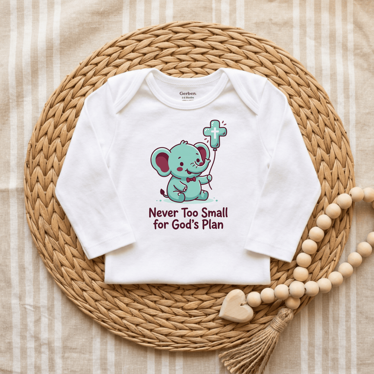 The Toodleroos Never Too Small for God’s Plan Onesie – Faith Bodysuit features a white baby onesie with a cartoon elephant holding a cross and the uplifting message “Never Too Small for God’s Plan.” Displayed on a woven placemat with a beaded heart decoration nearby, this Christian bodysuit makes a thoughtful baptism gift and is ideal for faith-inspired baby clothing collections.