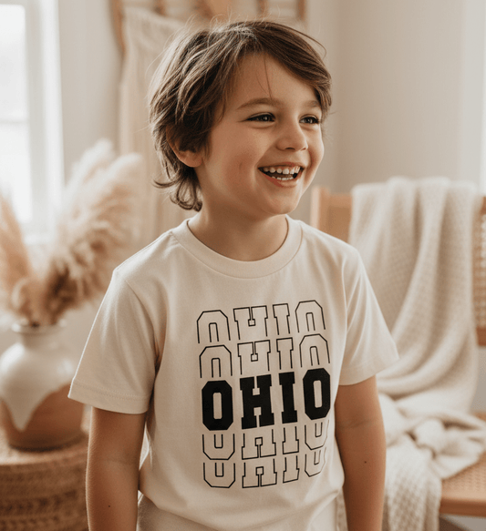 A smiling child with medium-length brown hair wears the Toodleroos Ohio Varsity Kids Shirt – State Pride Tee, featuring ‘OHIO’ three times in bold across the front, and stands in a cozy, softly-lit room adorned with neutral decor and pampas grass—a perfect tribute to Ohio pride.