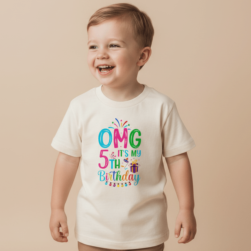 A smiling young child wears the Toodleroos OMG It’s My 5th Birthday Kids Shirt – Party Tee, a cream-colored shirt featuring playful, colorful letters spelling “OMG it’s my 5th Birthday,” accented with confetti and a small gift box graphic—the perfect birthday shirt for boys and girls.