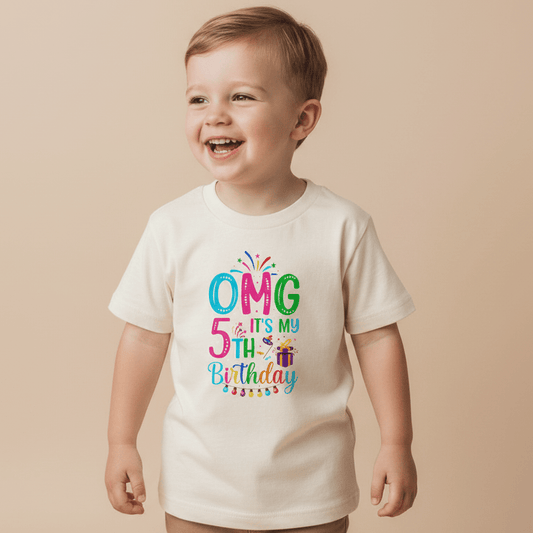 A smiling young child wears the Toodleroos OMG It’s My 5th Birthday Kids Shirt – Party Tee, a cream-colored shirt featuring playful, colorful letters spelling “OMG it’s my 5th Birthday,” accented with confetti and a small gift box graphic—the perfect birthday shirt for boys and girls.