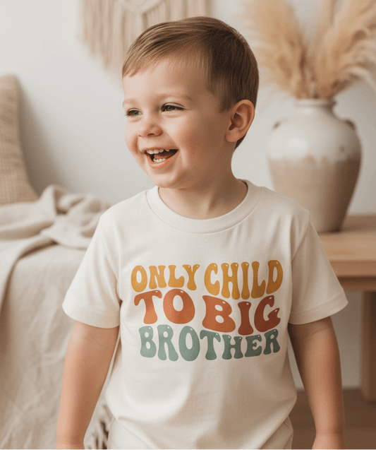 A smiling young boy wears the Toodleroos Only Child to Big Brother Toddler Shirt – Sibling Announcement Tee, standing indoors with neutral decor and a vase of pampas grass in the background—making it the perfect sibling reveal shirt for a sweet pregnancy announcement.