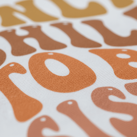 Close-up of textured fabric showcasing large, retro-style letters in warm brown and orange tones with rounded edges and a playful design on a white background—an ideal choice for the Toodleroos Only Child to Big Sister Toddler Shirt, perfect for sibling announcements or a big sister pregnancy reveal.