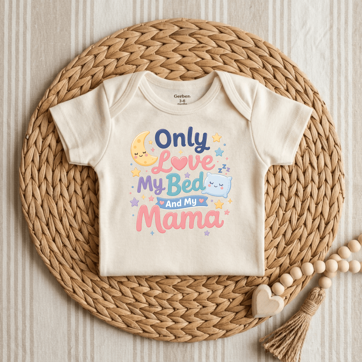 A cream-colored Toodleroos “Only Love My Bed and My Mama” Onesie rests on a woven mat, featuring colorful text, stars, a crescent moon, and a cheerful pillow graphic—making this cute gender-neutral baby bodysuit a perfect gift for anyone who loves funny onesies.