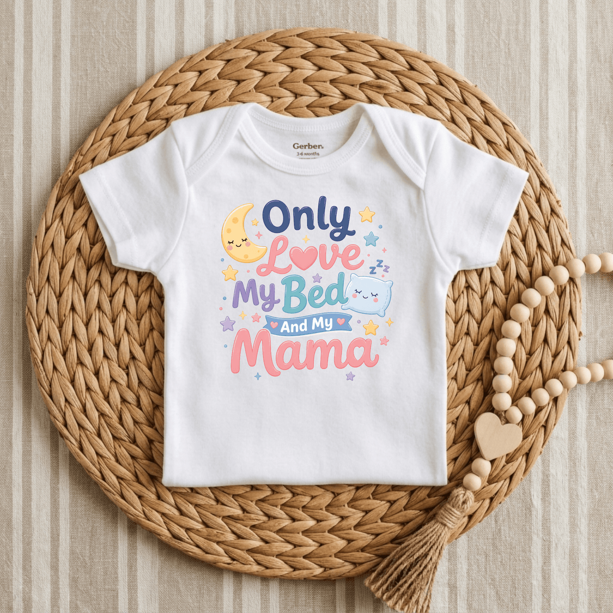 A Toodleroos “Only Love My Bed and My Mama Onesie – Cute Baby Bodysuit” in white is displayed on a woven placemat, featuring playful moon, star, and pillow illustrations along with colorful stars. It’s paired with a wooden bead garland for a charming touch, making this gender-neutral bodysuit a perfect baby gift.