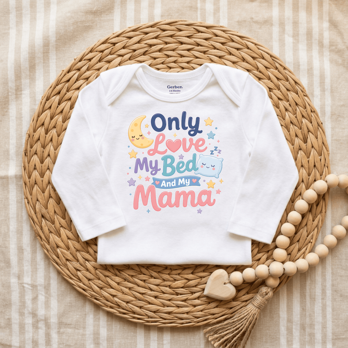 A Toodleroos Only Love My Bed and My Mama Onesie – Cute Baby Bodysuit in white, featuring colorful text, playful stars, a moon, and a pillow design, is displayed on a woven mat beside a beaded tassel with a heart—this funny, gender-neutral baby gift is perfect for any little one.