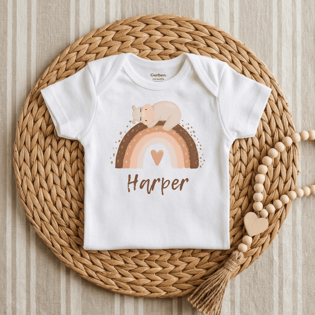 The Toodleroos Personalized Bear Rainbow Baby Onesie – Custom Name Bodysuit features a pastel rainbow and sleeping bear graphic with “Harper” printed below. Displayed on a round woven placemat beside a beaded tassel decoration, this charming, customized baby onesie makes the perfect boho baby gift.