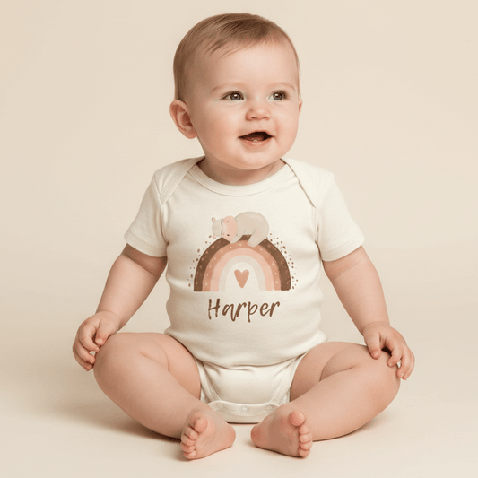 A smiling baby sits on a light background, wearing the Toodleroos Personalized Bear Rainbow Baby Onesie – a custom name bodysuit adorned with cheerful heart and bear designs. This boho-inspired onesie, personalized here with the name Harper, makes a wonderful baby gift.