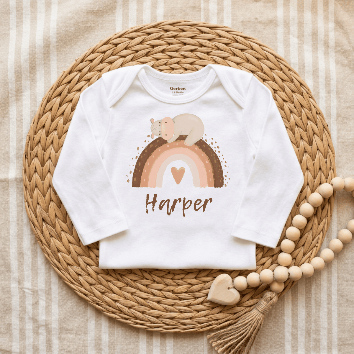 The Toodleroos Personalized Bear Rainbow Baby Onesie – Custom Name Bodysuit features a sweet heart and sleeping bear design accented with a charming rainbow. Custom printed with the name Harper, this white onesie is displayed on a woven mat beside a wooden bead garland, making it the perfect boho baby gift.