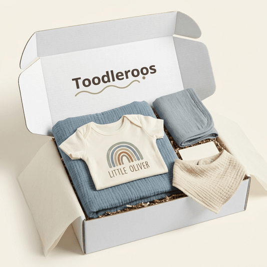 The Personalized Baby Gift Box – Boho Rainbow Custom Name Baby Shower Gift by Toodleroos features a beige boho rainbow baby onesie personalized with a rainbow and the name Little Oliver, a soft blue blanket, and a cream-colored bib, all beautifully presented on shredded paper.