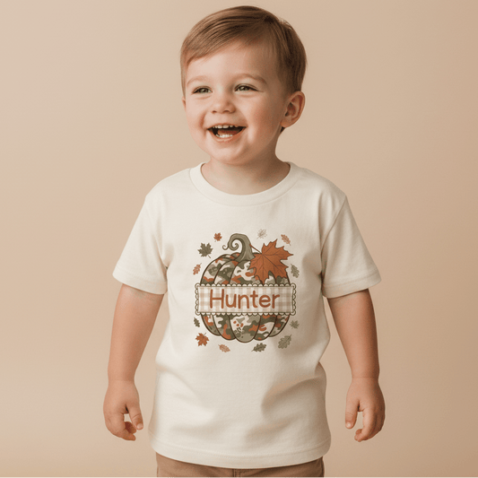 A smiling young child wears the Toodleroos Personalized Camo Pumpkin Toddler Shirt – Fall Name Tee, featuring a camo pumpkin, autumn leaves, and “Hunter” printed on the front, standing against a beige background.