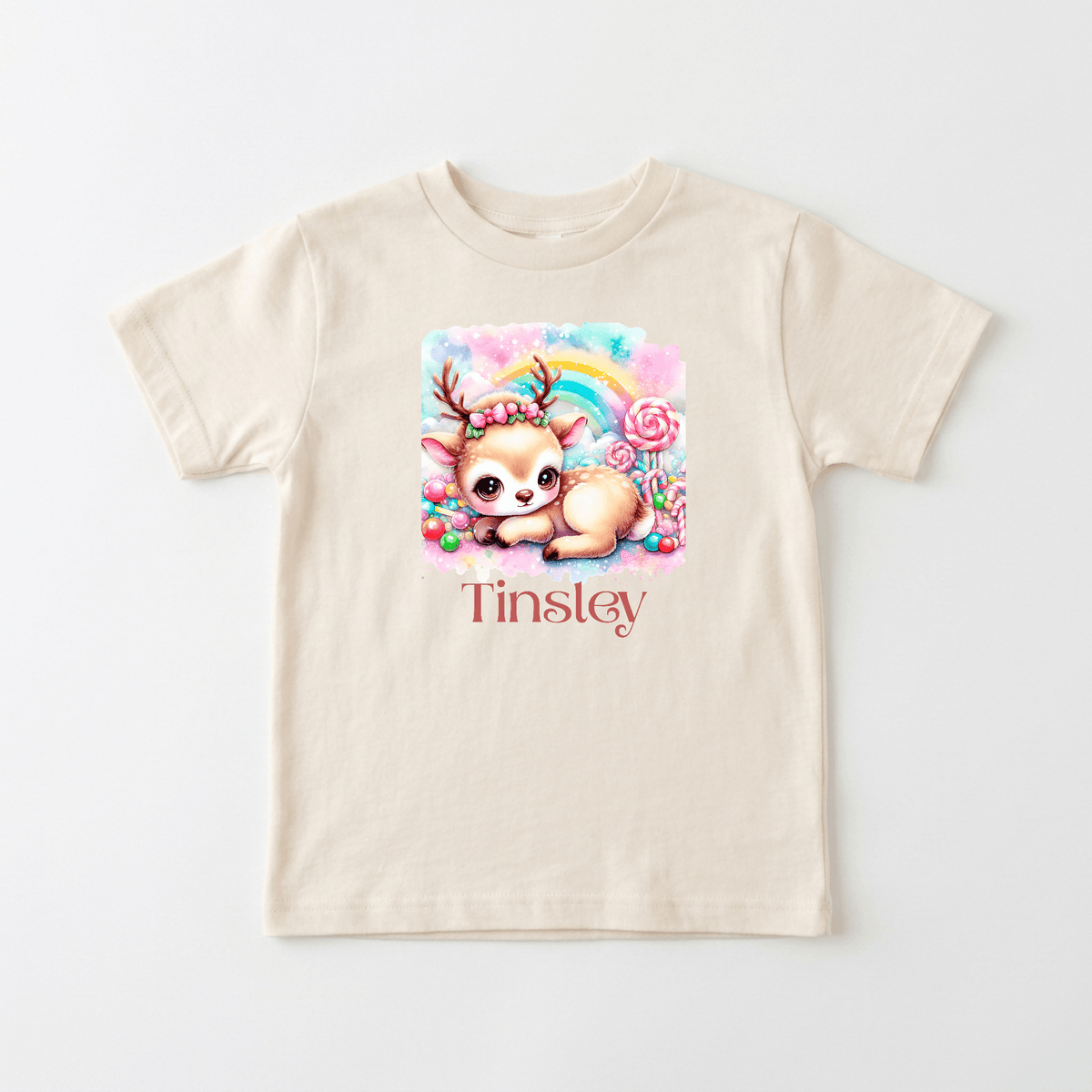 The Toodleroos Personalized Candy Deer Toddler T-Shirt – Custom Name Tee features a cream-colored design with a charming cartoon baby deer, sweet candy accents, and a pastel rainbow graphic above your child’s name in bold red letters. Bright and playful, this customizable t-shirt is perfect for toddlers.