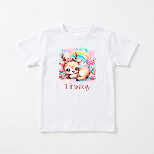 Introducing the Personalized Candy Deer Toddler T-Shirt – Custom Name Tee by Toodleroos! This adorable white t-shirt features a cute cartoon baby deer, colorful candy, and a pastel rainbow. Your child’s name, such as “Tinsley,” is printed in bold red beneath the illustration for a charming personalized touch—making it the perfect toddler tee.