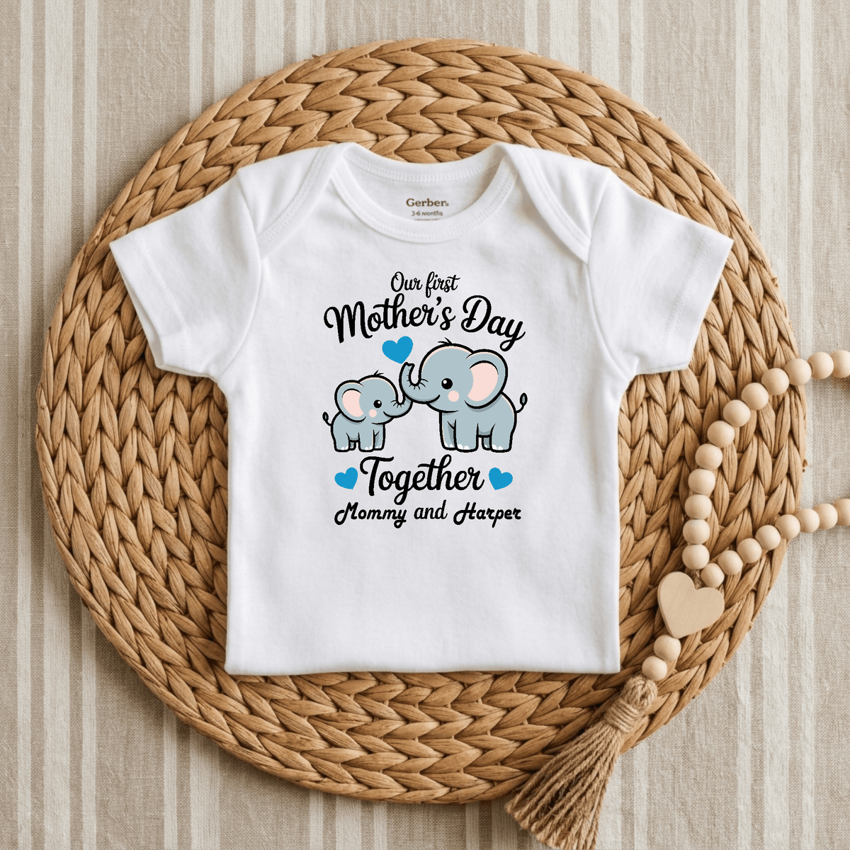A Personalized First Mother’s Day Onesie – Custom Baby Bodysuit by Toodleroos rests atop a round woven placemat. This adorable Mother’s Day outfit is adorned with an illustration of a mommy and baby elephant with blue hearts, along with the text: “Our first Mothers Day Together Mommy and Harper.” Wooden beads accent the placemat, making this Toodleroos personalized onesie a delightful keepsake for mom and baby.