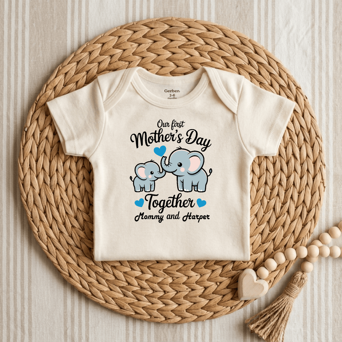 A Toodleroos Personalized First Mother’s Day Onesie – Custom Baby Bodysuit is displayed on a woven mat, decorated with two elephants and blue hearts, and the phrase “Our first Mothers Day together Mommy and Harper.” The Gerber short-sleeve bodysuit is shown alongside a wooden teething toy.