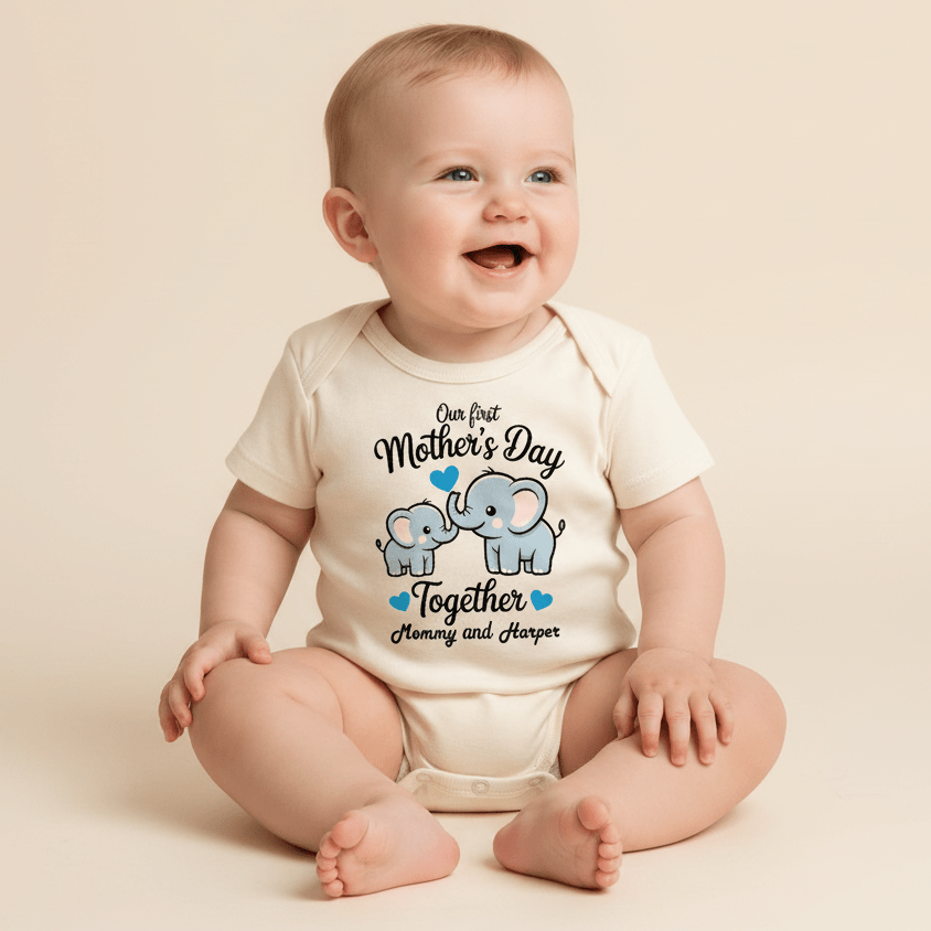 A smiling baby sits wearing a cream Toodleroos Personalized First Mother’s Day Onesie – Custom Baby Bodysuit, featuring a charming design of two elephants with blue hearts and the message “Our first Mothers Day together, Mommy and Harper”—the perfect personalized gift for celebrating a first Mother’s Day.