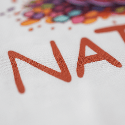 Close-up of red, textured letters “NAT” printed on white fabric, with colorful abstract shapes and dots above the text—a perfect design for the Toodleroos Personalized Ghost Pumpkin Toddler Shirt – Halloween Name Tee.