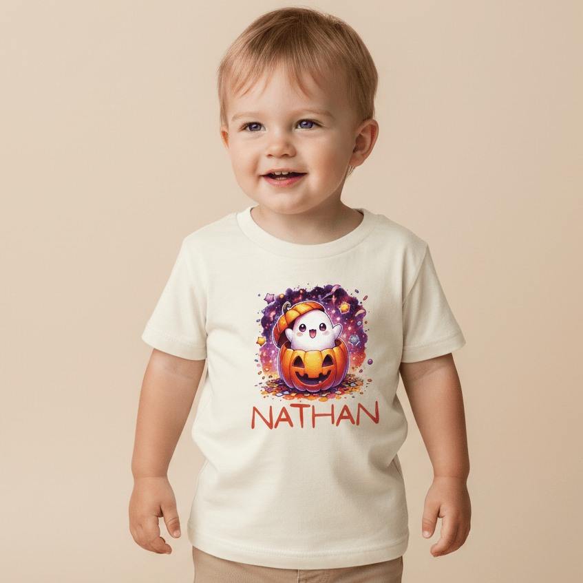 A happy toddler wears the Toodleroos Personalized Ghost Pumpkin Toddler Shirt – Halloween Name Tee, featuring a cute ghost inside a pumpkin and the name Nathan printed below in red letters—an adorable custom Halloween shirt.