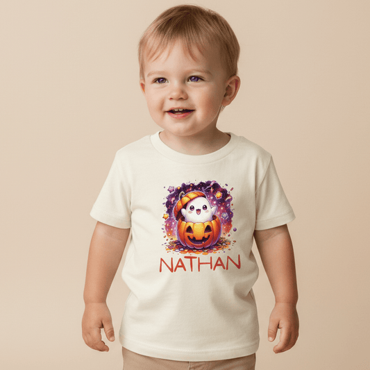 A happy toddler wears the Toodleroos Personalized Ghost Pumpkin Toddler Shirt – Halloween Name Tee, featuring a cute ghost inside a pumpkin and the name Nathan printed below in red letters—an adorable custom Halloween shirt.