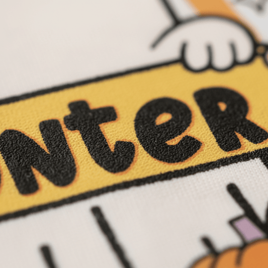 Close-up of printed text on fabric displaying bold, black letters outlined in orange on a yellow background, with part of a cartoon hand above and a pumpkin below—featured on the Toodleroos Personalized Halloween Ghost Kids Shirt – Custom Name Tee.