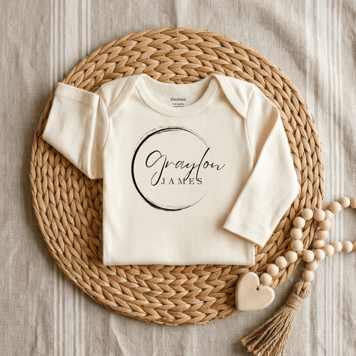 A cream-colored Personalized Name Onesie – Custom Baby Bodysuit by Toodleroos, featuring “Graydon James” written in script inside a black circle, displayed on a woven mat beside a beaded tassel with a wooden heart.