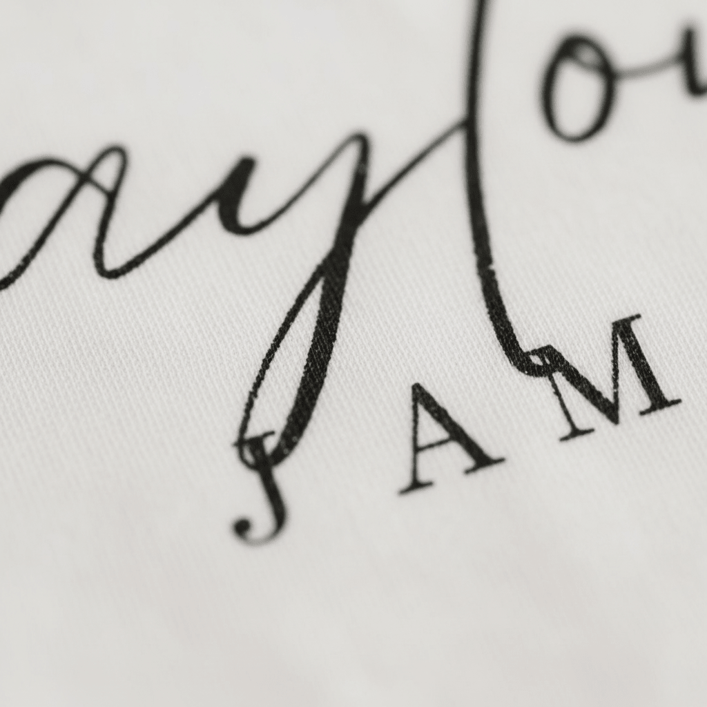 A detailed close-up showcases black cursive and uppercase printed text reading the partial name “Taylor” and the word “JAM” on crisp white fabric. The image highlights the fine texture and expert craftsmanship of Toodleroos’ Personalized Name Onesie – Custom Baby Bodysuit, capturing its high-quality custom printing.